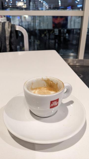 a small cup of cafe machiato, white ily branded espresso cup, white table, a dark, hazy, rainy tarmack is in the background.