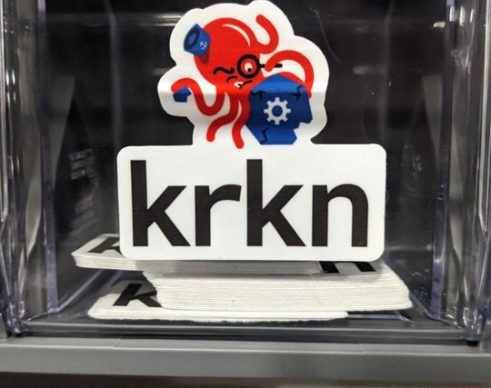 sticker with a red octopus wearing a blue hat and holding a blue kubernetes logo, large text reads "krkn"