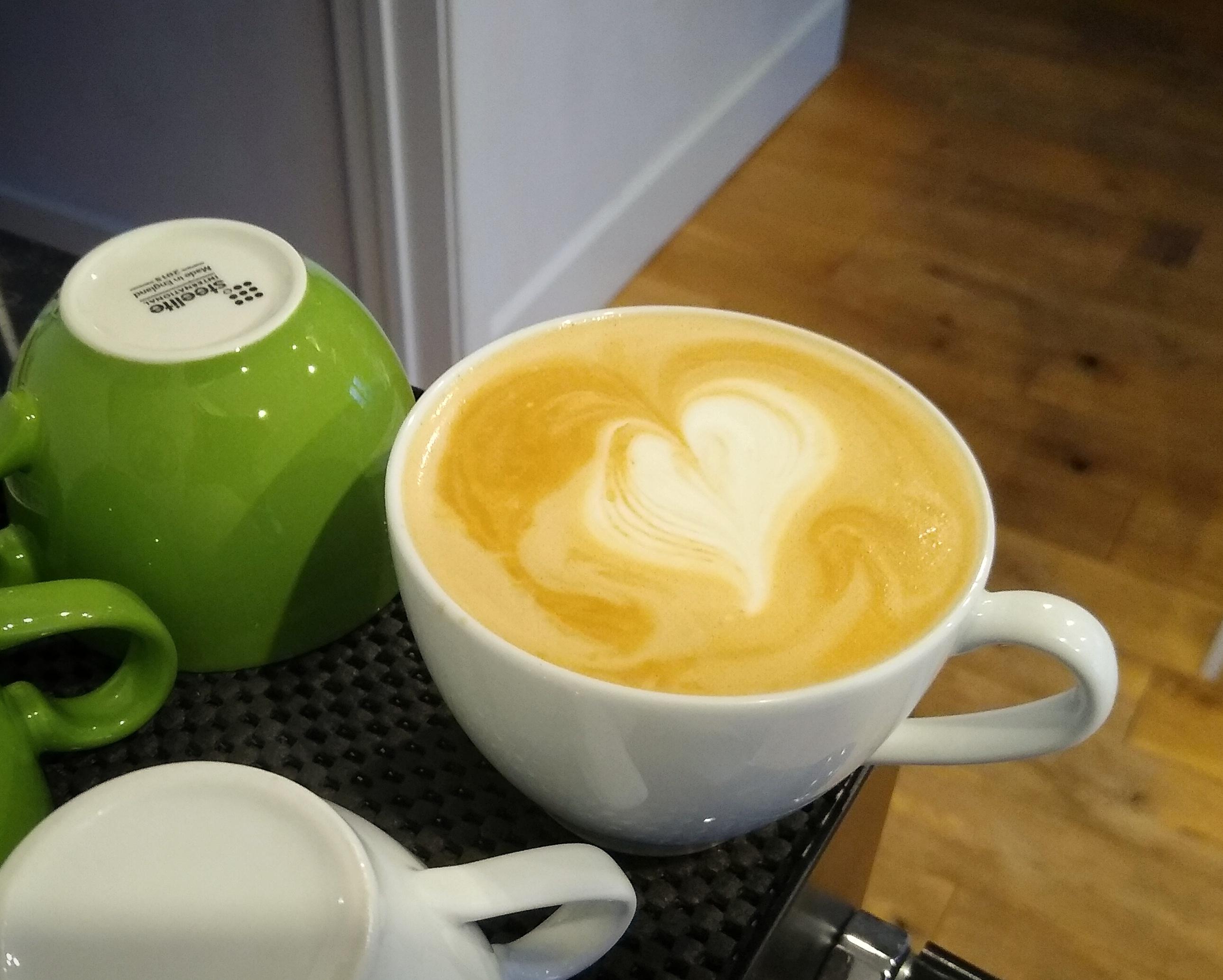 A coffee cup, with a slightly off-kilter heart shape pattern in the milk.