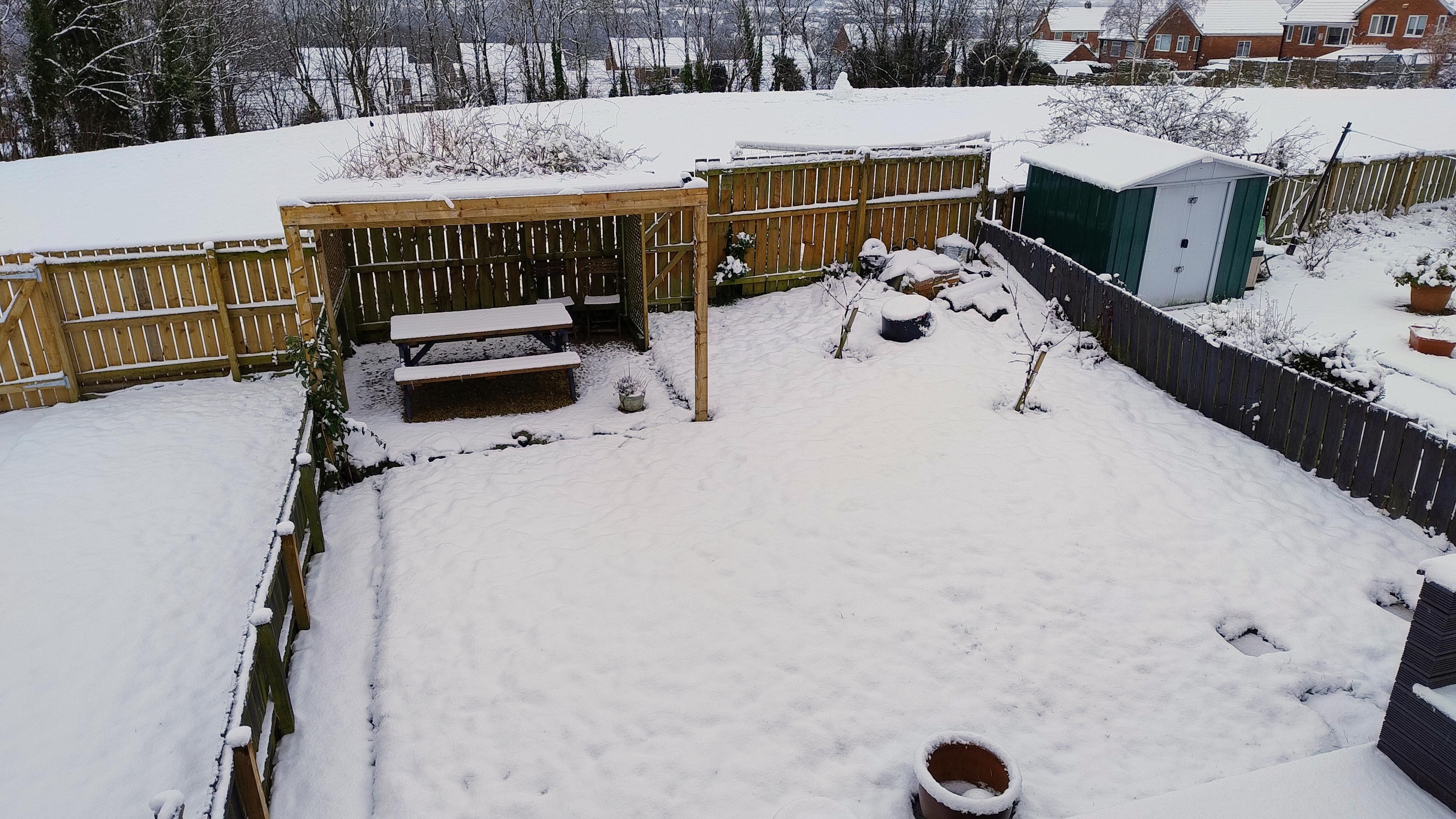 Snowy suburban back garden. Snow covering what is probably grass, and layers of snow over a picnic bench and wooden arbour structure.