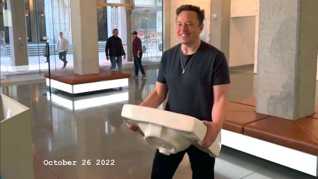 A man enters what appears to be the ground floor of an office building with a sink in his hands. The man wears a black t-shirt and black trousers. On the bottom left a date is written: October 26 2002. The man is smiling.