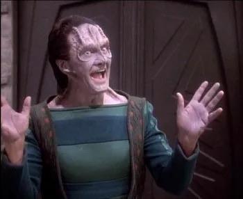 A Cardiassian, Garak, featuring a grayish skin tone and forehead ridges, expresses glee at the latest Cardiassian novel while gesturing widely with both hands while speaking. 