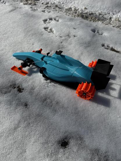 A photo of a blue and black OpenRC F1 car with orange skis replacing the front wheels and orange paddle wheels replacing the rear wheels. The car is sitting on a snowy driveway.