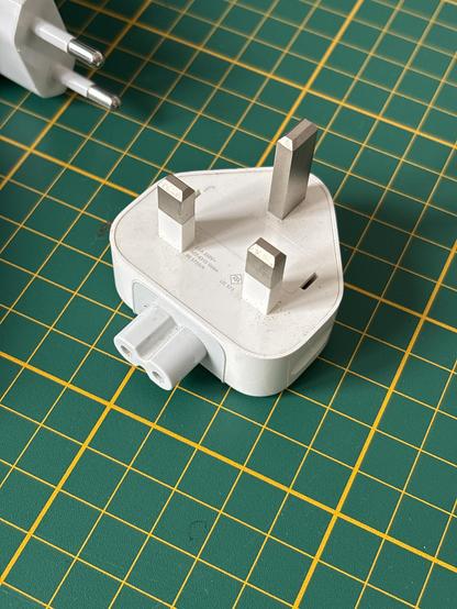 Image of a white three pronged plug, of the kind they have in Ireland. There’s a European one showing in the corner. The background is a green cutting mat with a grid pattern.