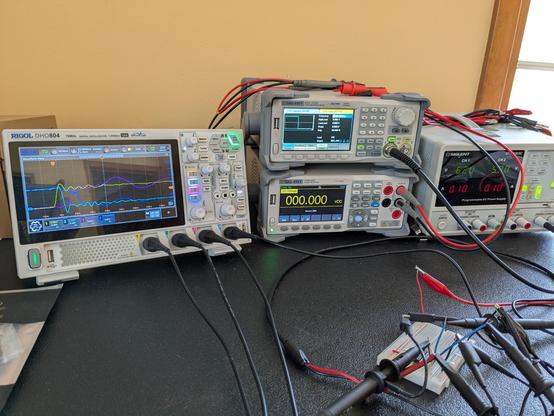 Electronics lab bench with oscilloscope, signal generator, and power supply with leads connected to a small breadboard circuit.