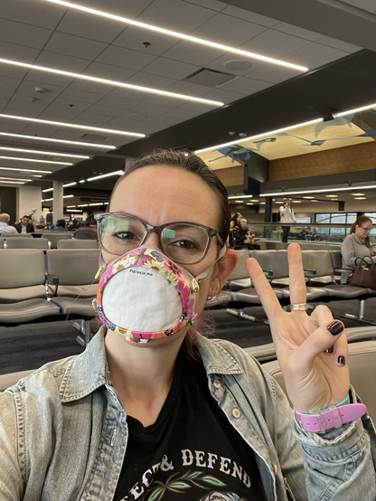 Me (40 y/o white brunette) wearing glasses, a special edition floral Flo mask, and a Protect and Defend Trans Kids tee and giving a peace sign from a seat at my gate at Pittsburgh airport 