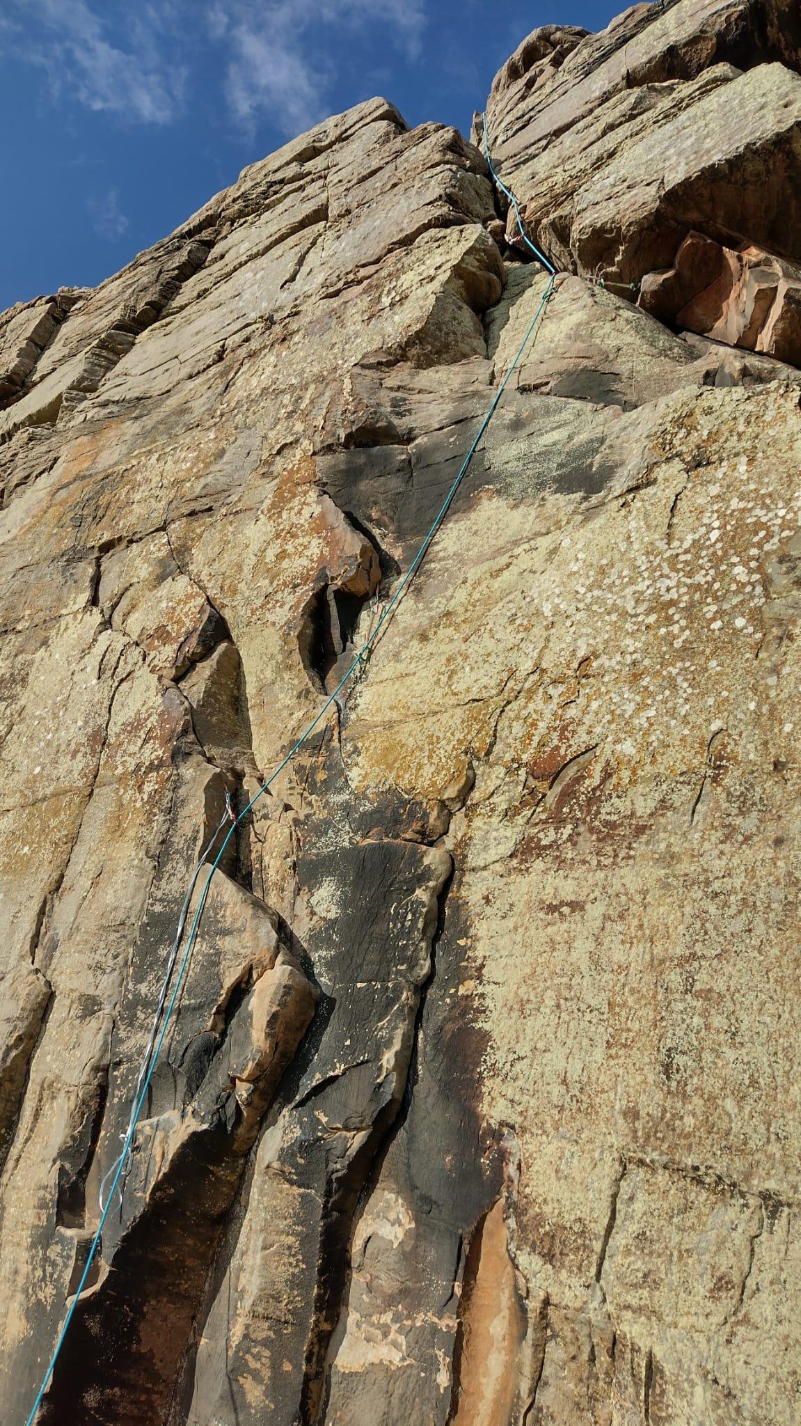 A sandstone crag beneath blue skies. A rope with a few runners is trailing upwards following a crack. (Why is there a sling dangling from the first nut? My friend was worried she might not be able to reach the first hold, but actually she was fine! 👍)