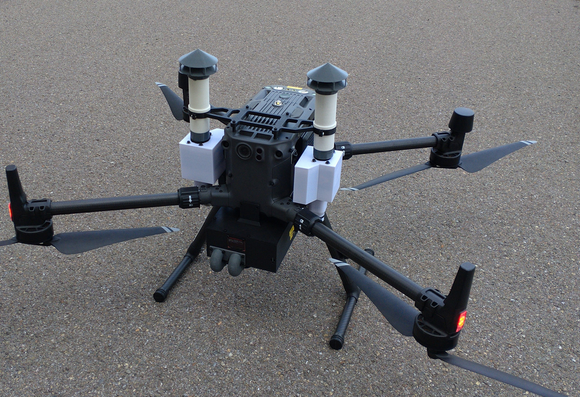 A dji Matrice M300 multicopter on asphalt with 3d printed parts attached all over, two "chimineys" at either side for particle measurements and a black "belly" underneath, with temperature, humidity and gas sensors.