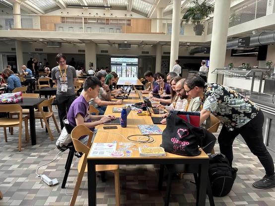 Django table at the EuroPythons prints, with 11 participants around the table focusing on their laptops, other sprint participants in the background, django girls tote bag in the foreground