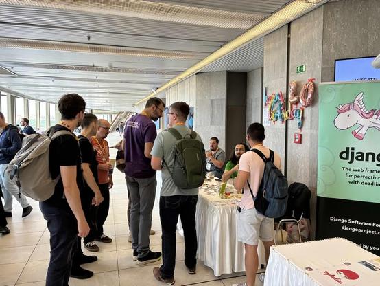 7 conference attendees at the django booth, talking to two volunteers behind the booth, the booth has balloons saying 'happy birthday 20', and a banner with the django pony