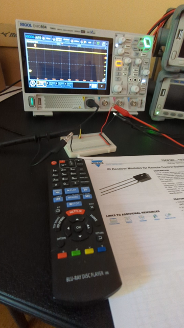 A TV/Blu-Ray player remote control pointed at a simple circuit on a proto-board. When buttons are pressed on the remote, the oscilloscope in the background displays pulse-width modulated patterns. Next to the remote is a datasheet for the IR receiver module on the proto-board.