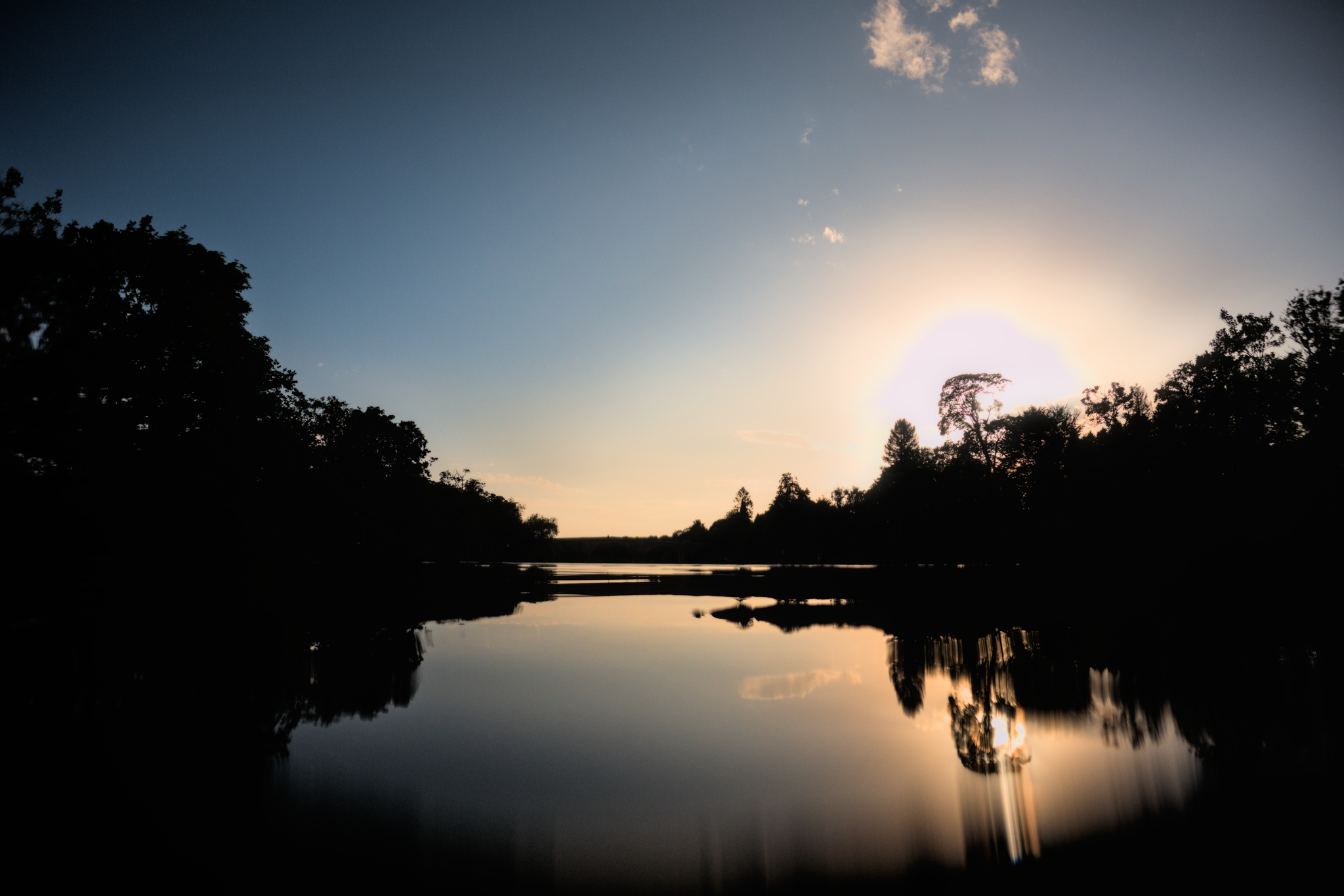 GoPro photo from the middle of a calm river, looking towards the sun. Almost no clouds, and the exposure straight into the sun casts the trees (and their reflections) in silhouette.