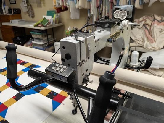 A longarm quilting machine with handlebar controls with thumb buttons. The right handlebar has wires sticking out of the end attached with black electrical tape to a push button switch that is way too small for the button enclosure. On the machine sewing table, there is a quilt with a white background and yellow, blue, red, and black squares and triangles.
