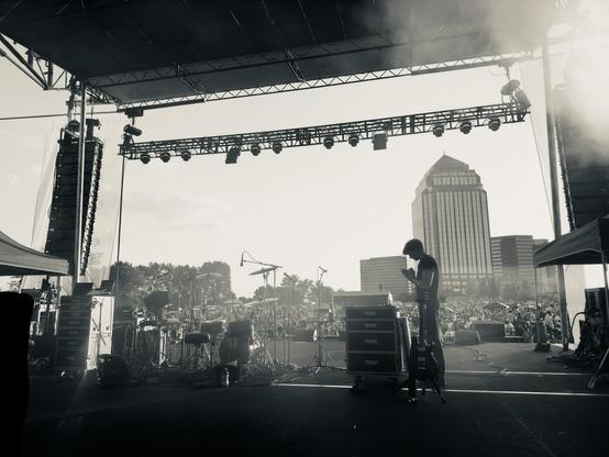 Moody black and white photo of a sound engineer standing on a festival stage checking the backline, looking at their iPad, with a crowd of thousands and some tall buildings in the background.