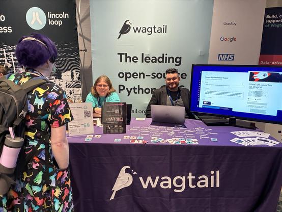Meagen and Vince at the Wagtail booth, with an attendee in a colorful dress in the foreground. Meagen and Vince are seating behind the booth table covered in stickers, with a Wagtail logo on the cloth, and a TV showcasing the admin interface