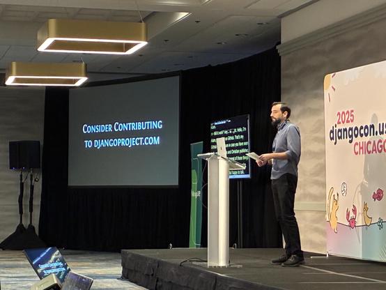 Adam on stage with a slide that says "Consider contributing to Djangoproject.com"