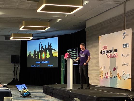 Tim on stage behind the lectern at DjangoCon US 2025 in Chicago, with a slide that says "What a decade!"