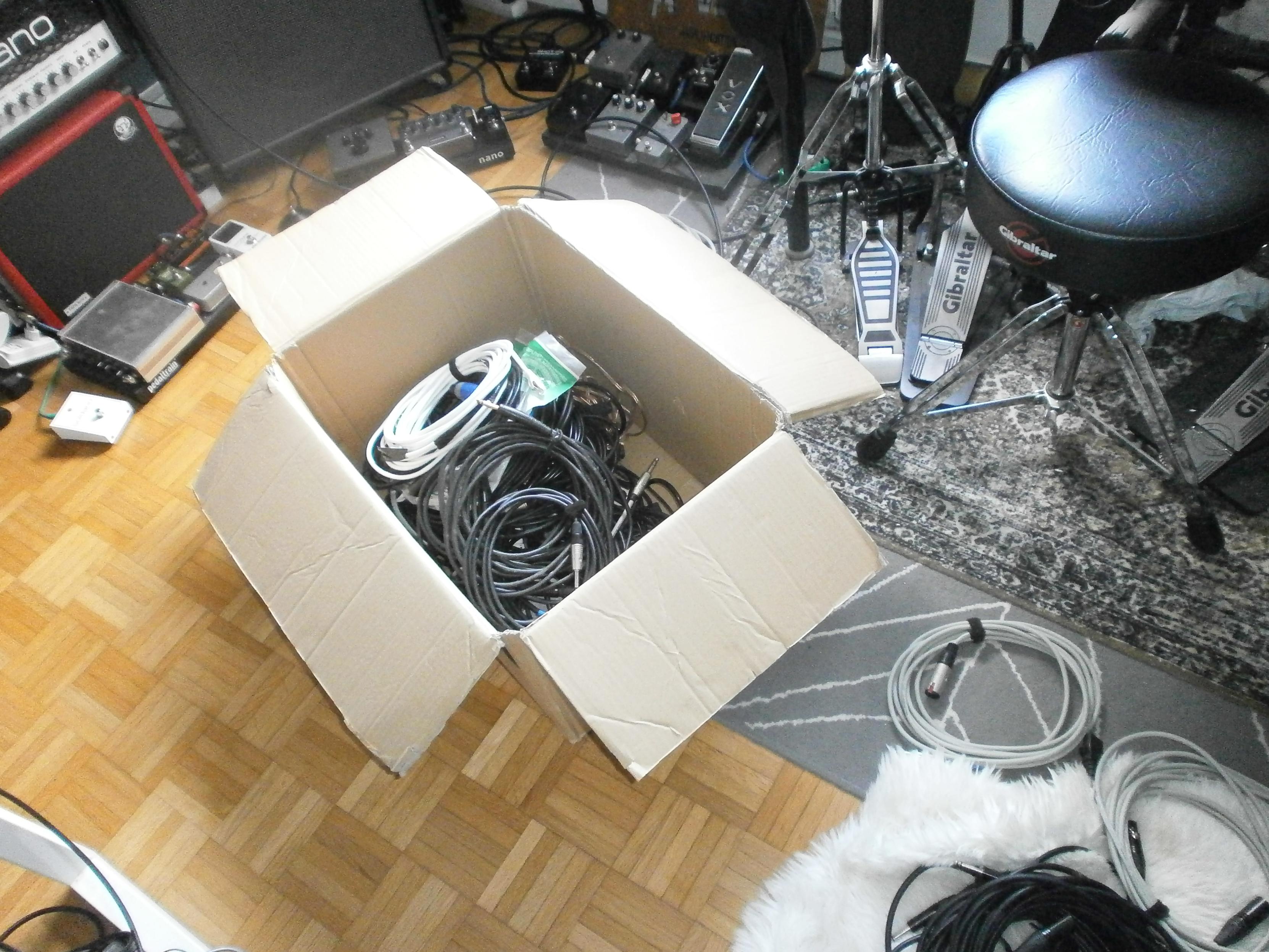 An open cardboard box containing lots of cables lies on the floor, with a few more cables next to it. Behind the box are a few amplifiers and pedalboards, and on the right one can see a drummer's seat and a hi-hat + kick drum pedals.