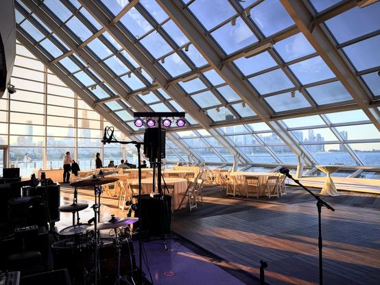 A view of the Chicago skyline from across the lake in the solarium of the Adler Planetarium. Instruments are silhouette in purple stage lights in the foreground, in the mid ground we see round event tables bathed in golden late afternoon sunlight.