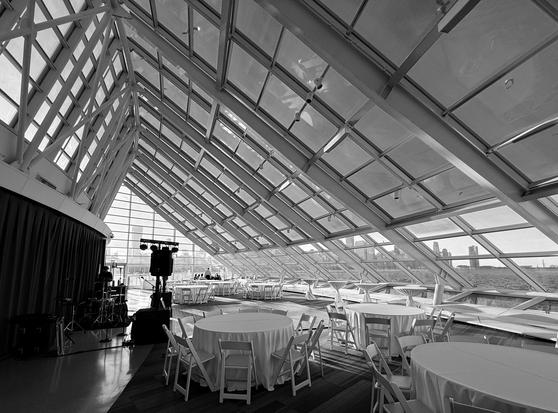 A black and white, wider angle photograph of the solarium at the Adler planetarium. The space is set up with special event round tables, and a band has set up on the left-hand side against the wall. Visible in the background through the glass is the Chicago skyline.