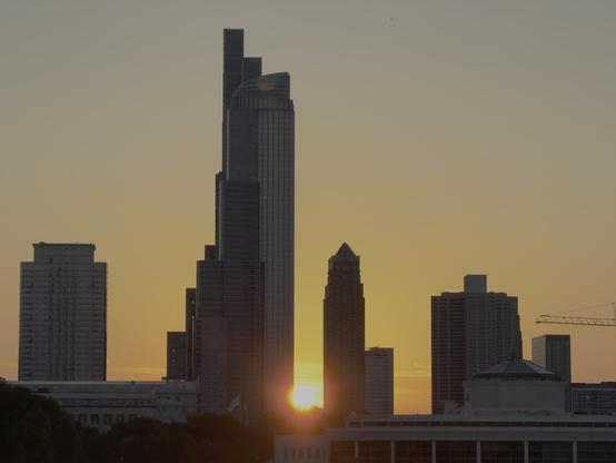 A handful of skyscrapers are silhouetted against the orange sky of the setting sun, the sun peeking through between two of them