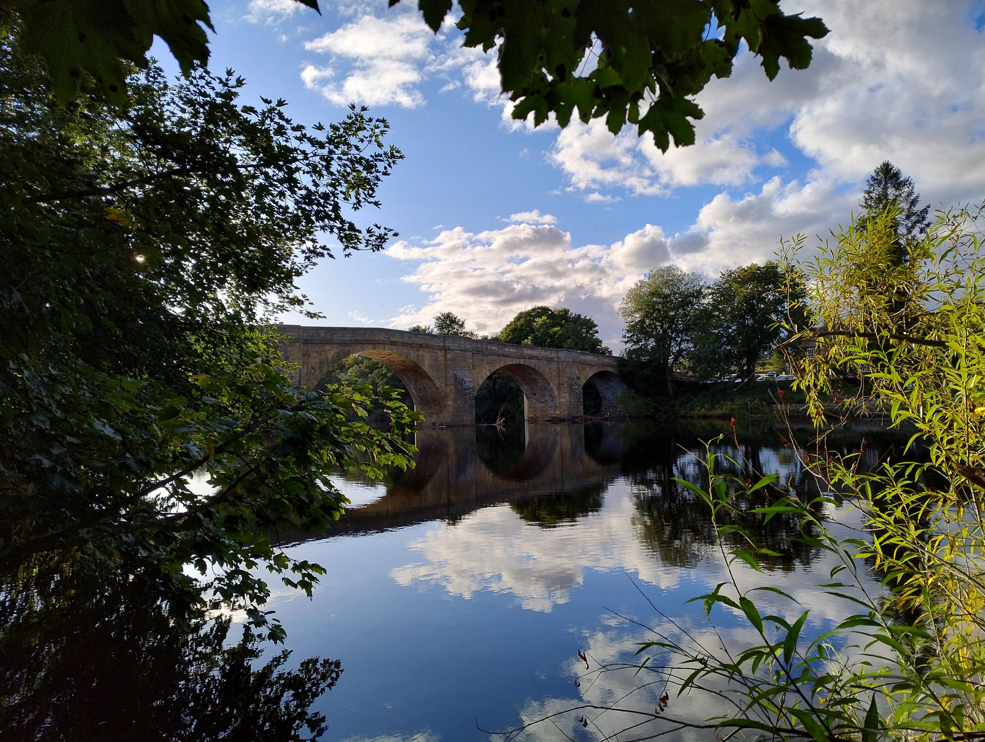 Photo of an old stone bridge seen from a river bank, framed with tree branches and sky reflected in calm water.