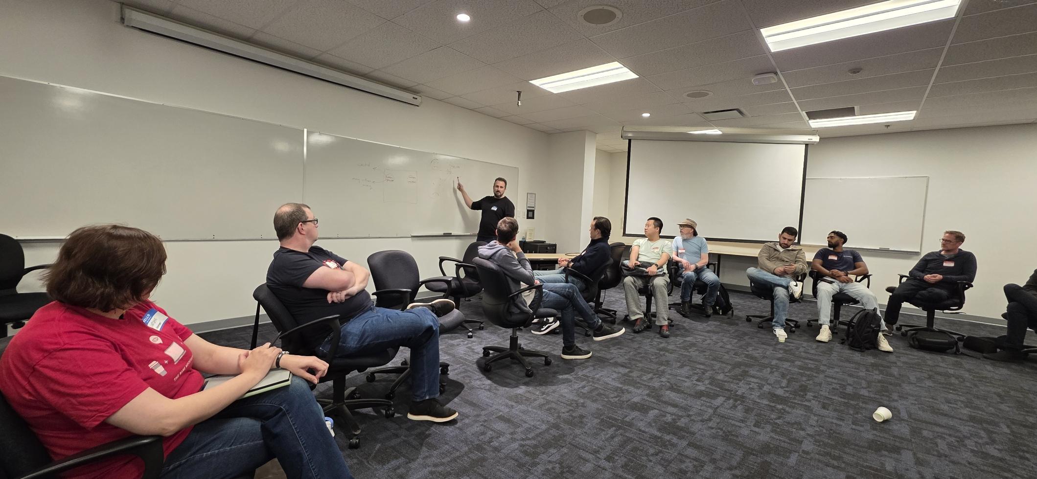 Participants seated in chairs, while one person is writing and pointing to the white board