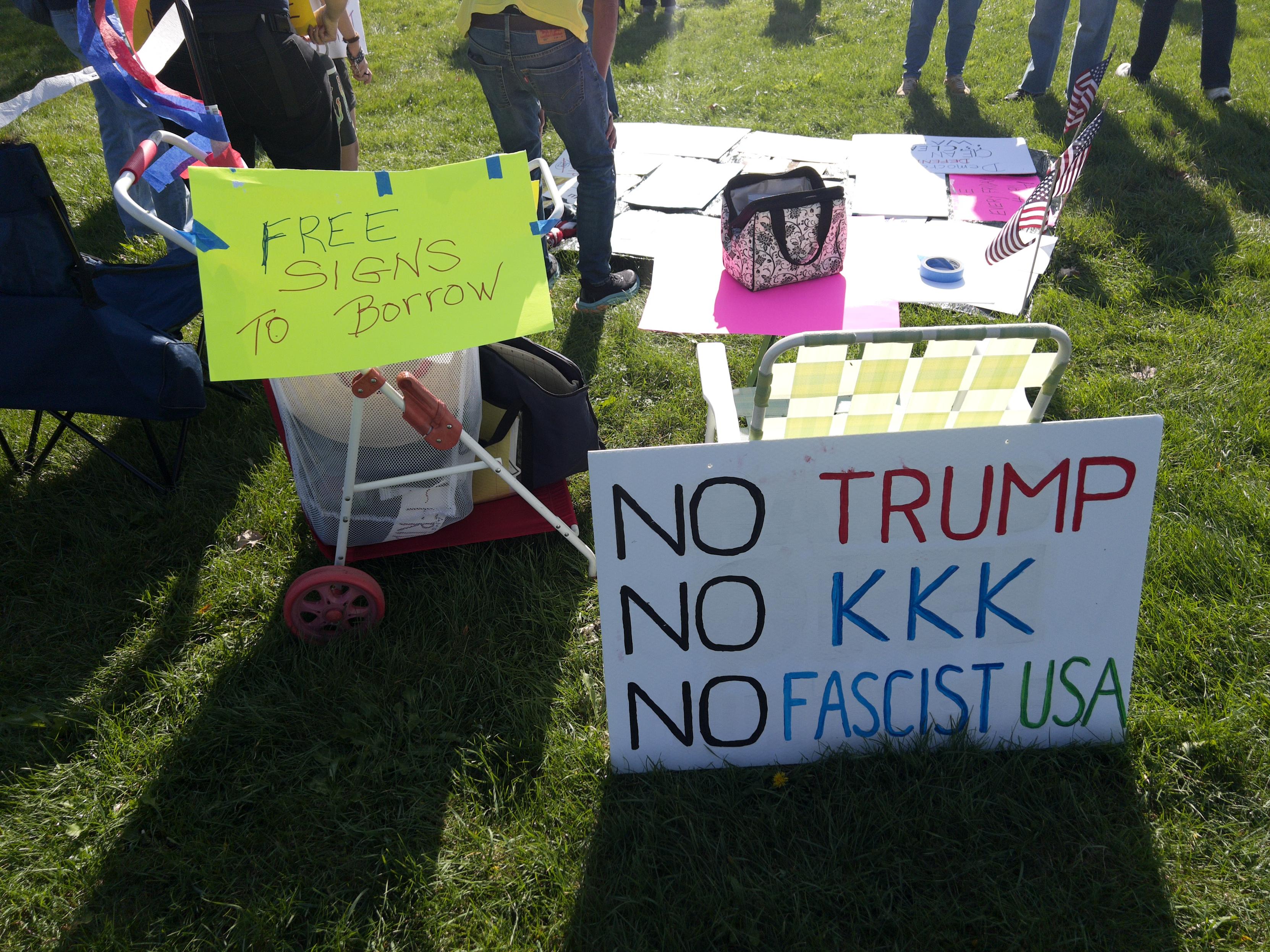 A cart with a sign reading “Free signs to borrow”. Another sign nearby reads “NO TRUMP / NO KKK / NO FASCIST USA”