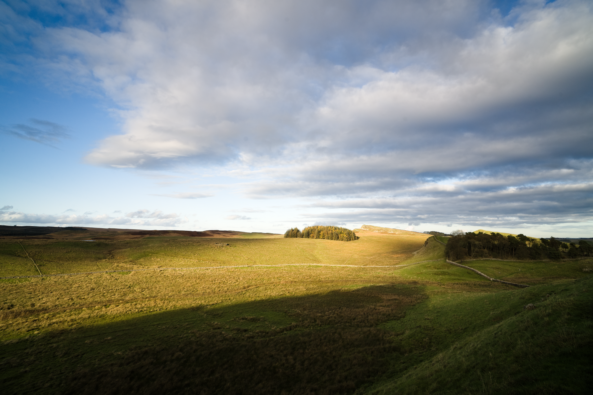 Open country landscape, mixed cloudy skies. Dark and shadowy foreground, but sunlight is catching the middle-distance, illuminating grass and trees.
