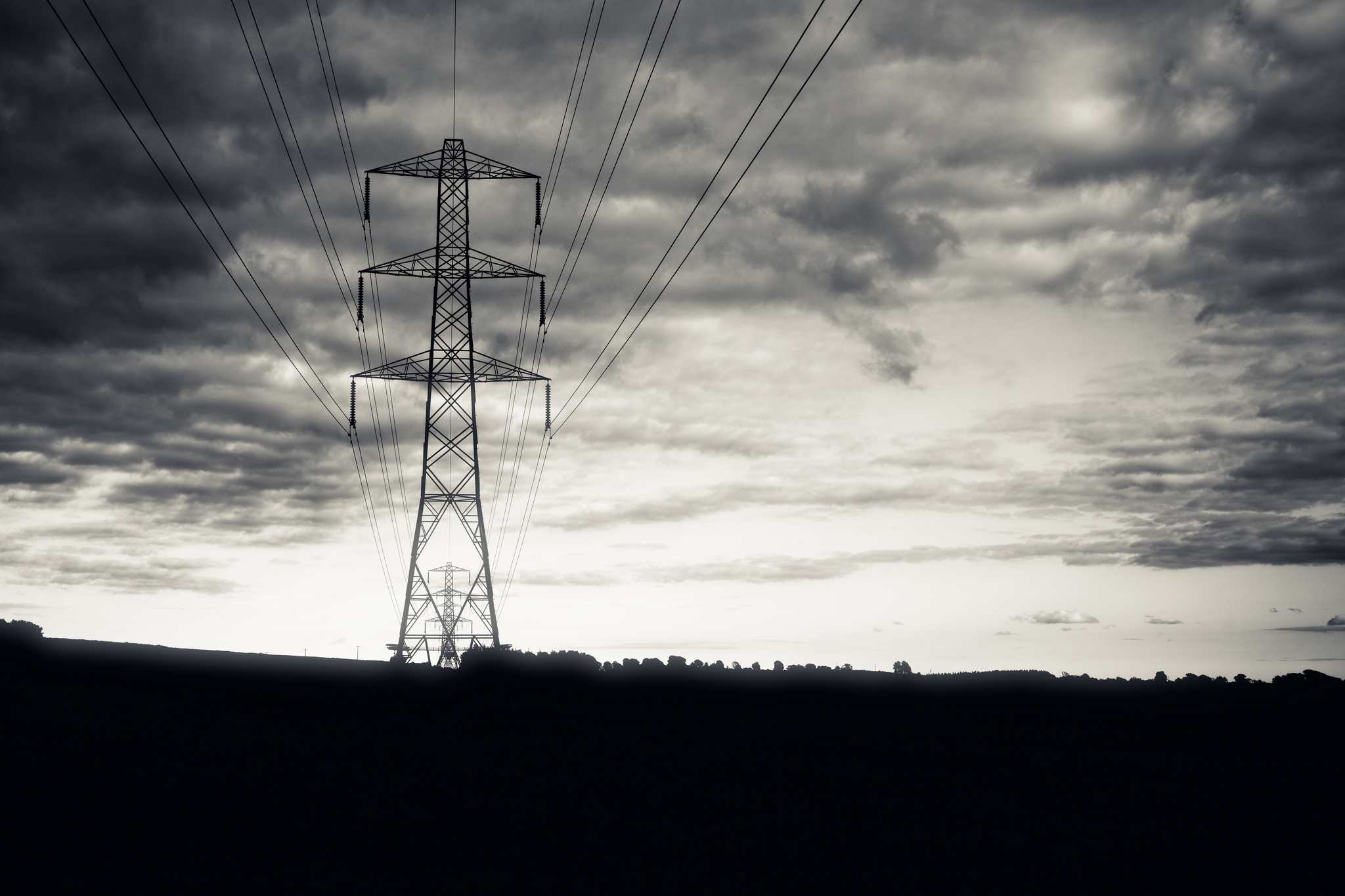 Monochrome image of an electricity pylon, in silhouette, against a cloudy sky.