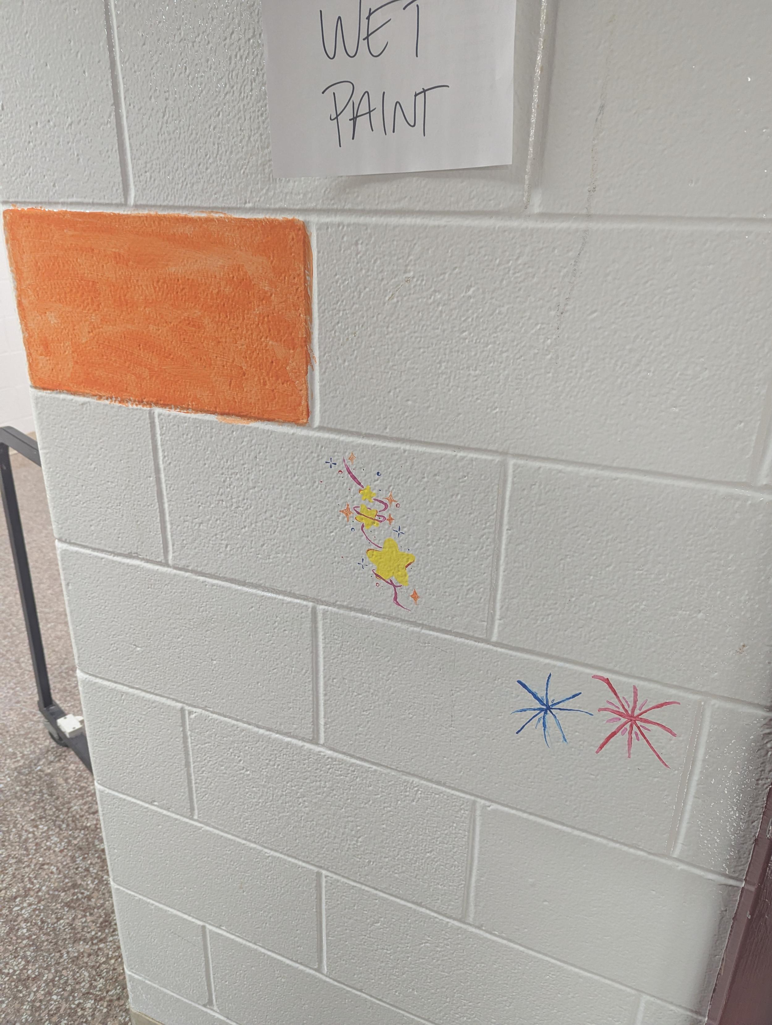 A white cinder block wall with three small murals in progress. A bright orange square is ready for detail work. Another block has stars showing technetium's presence in some special stars. A third has fireworks showing how cesium is used to color the displays.