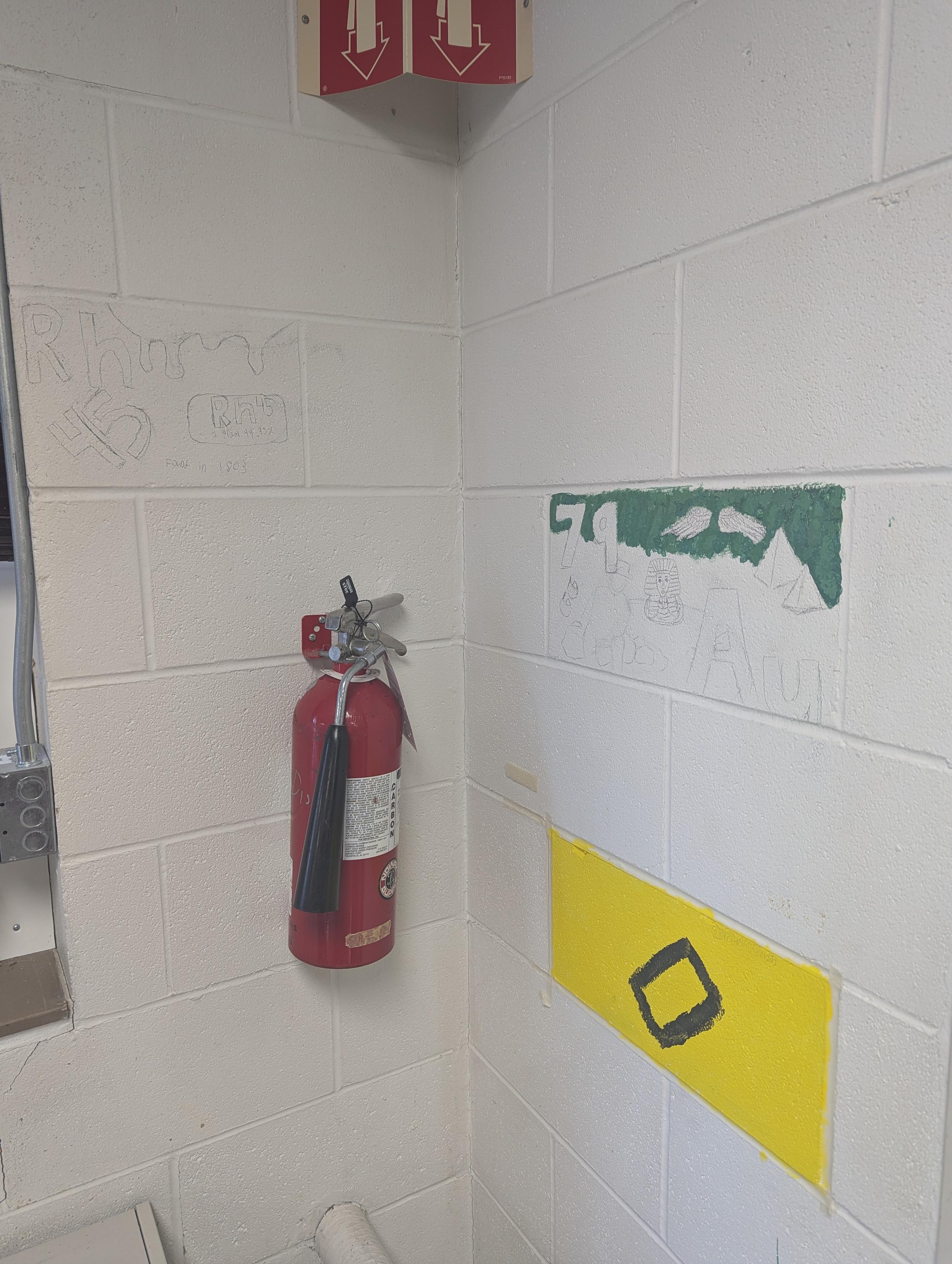 A white cinder block wall with three small murals about chemical elements in progress. One is bright yellow. Another has Egyptian imagery showing that gold has been used for thousands of years.