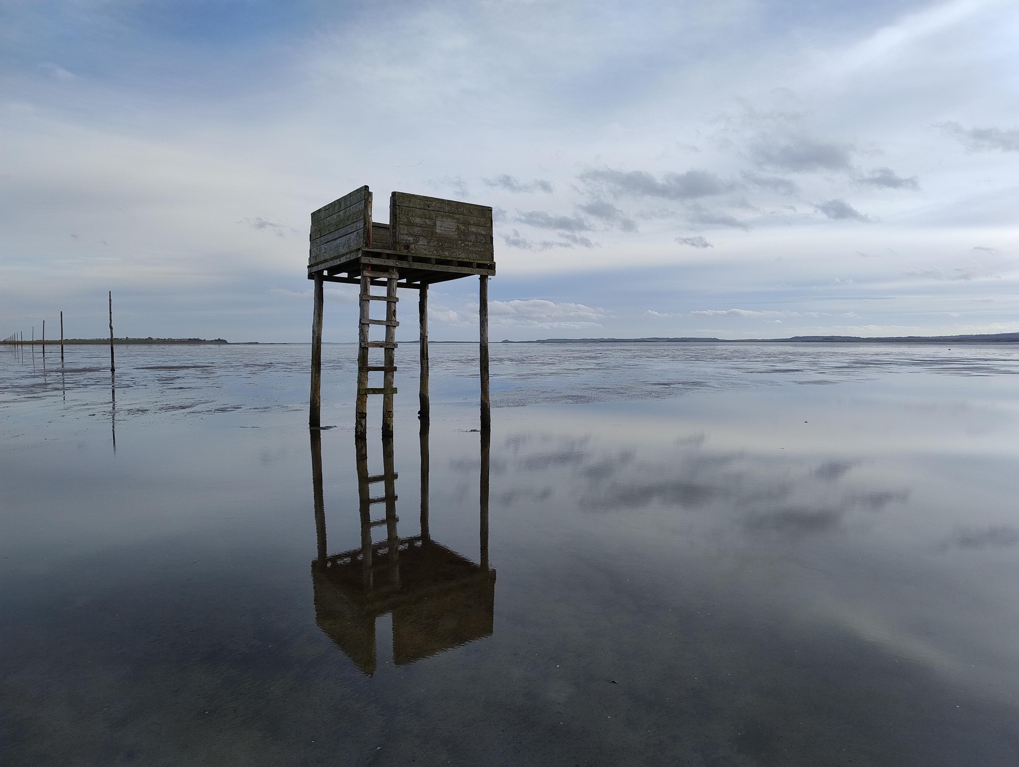 Emergency tide shelter, wooden platform on stilts, with Holy Island in the background. The shelter is mirrored in a pool.
