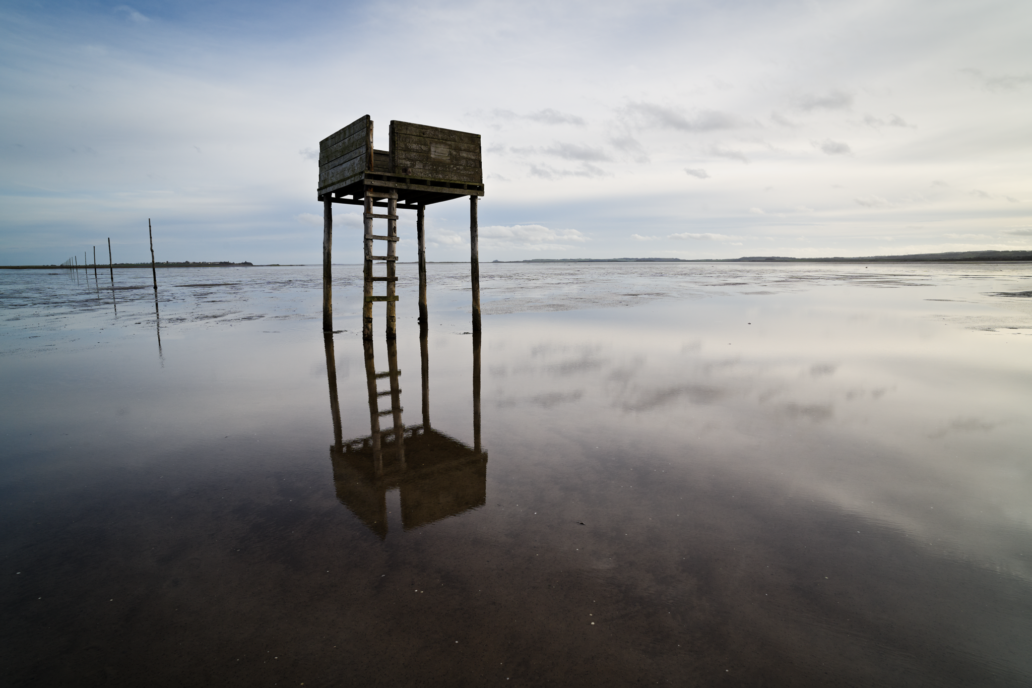 Emergency tide shelter, wooden platform on stilts, with Holy Island in the background. The shelter is mirrored in a pool.