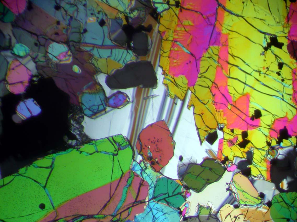 A thinsection imaged in cross-polarized light. Brightly colored pyroxene and olivine grains surround gray laminated plagioclase feldspar. field of view ~1mm. Sample 12005.