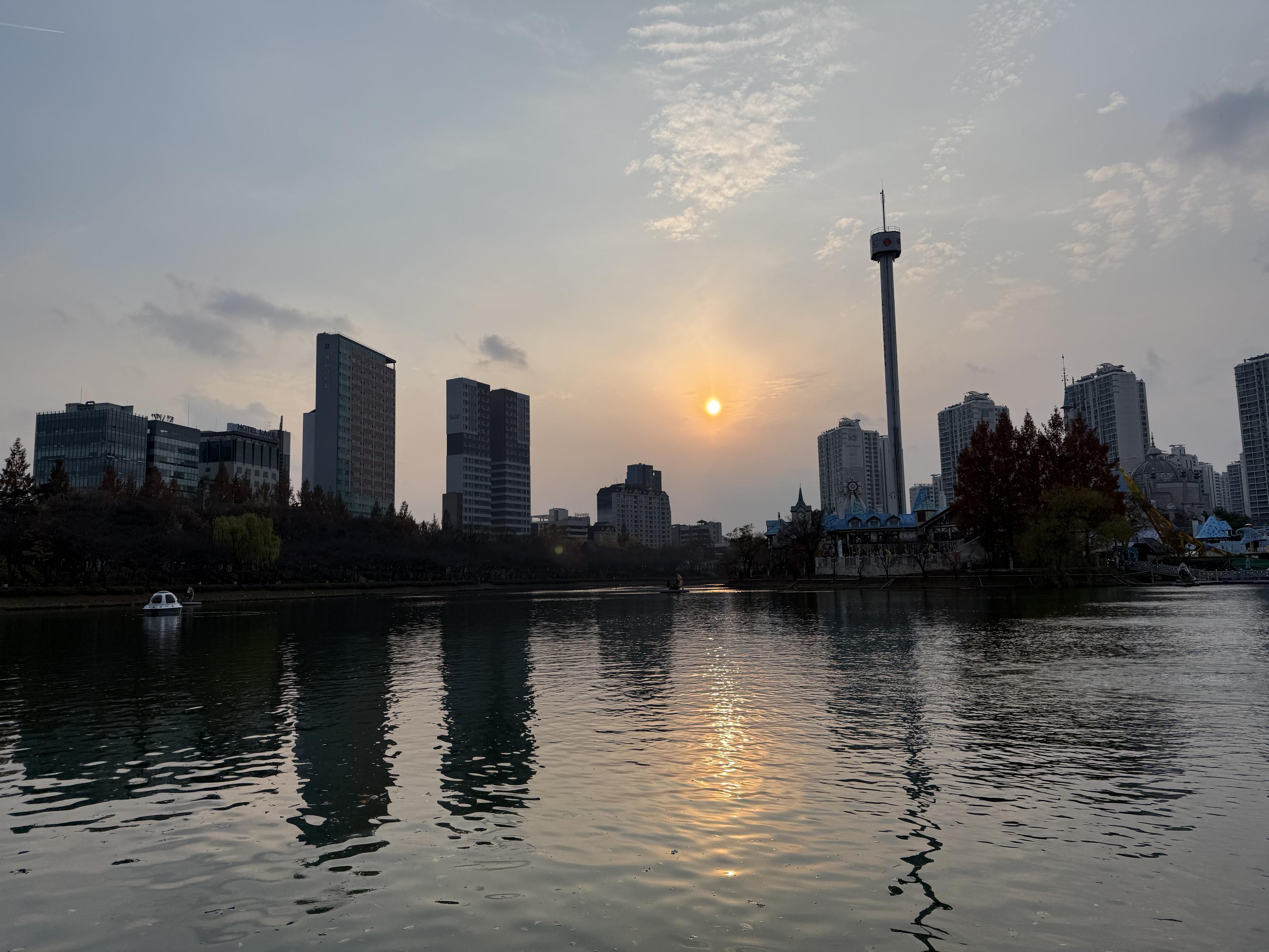 A lake in late afternoon with the sun low on the horizon. Several apartment and office buildings are visible around the edges. The buildings and a tower have skewed reflections due to ripples on the surface of the water.