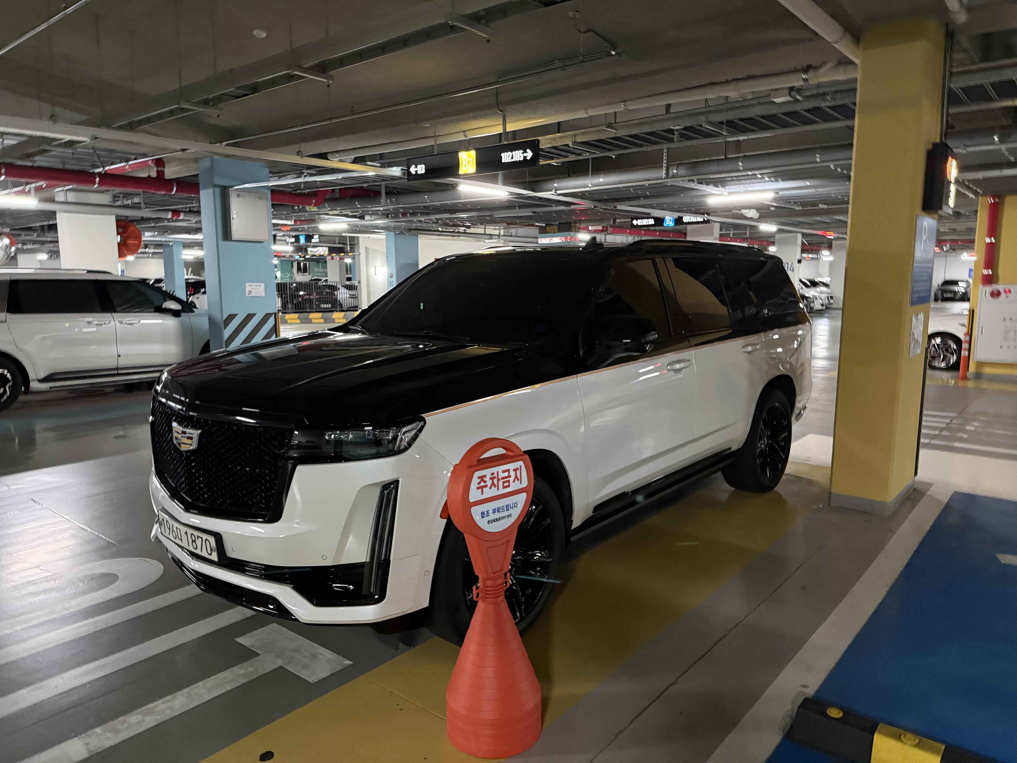 A Cadillac Escalade SUV with the entire upper section solid black. Illegally parked partially blocking traffic flow in a parking garage. Ironically directly in front of a sign that says “no parking” in Korean.