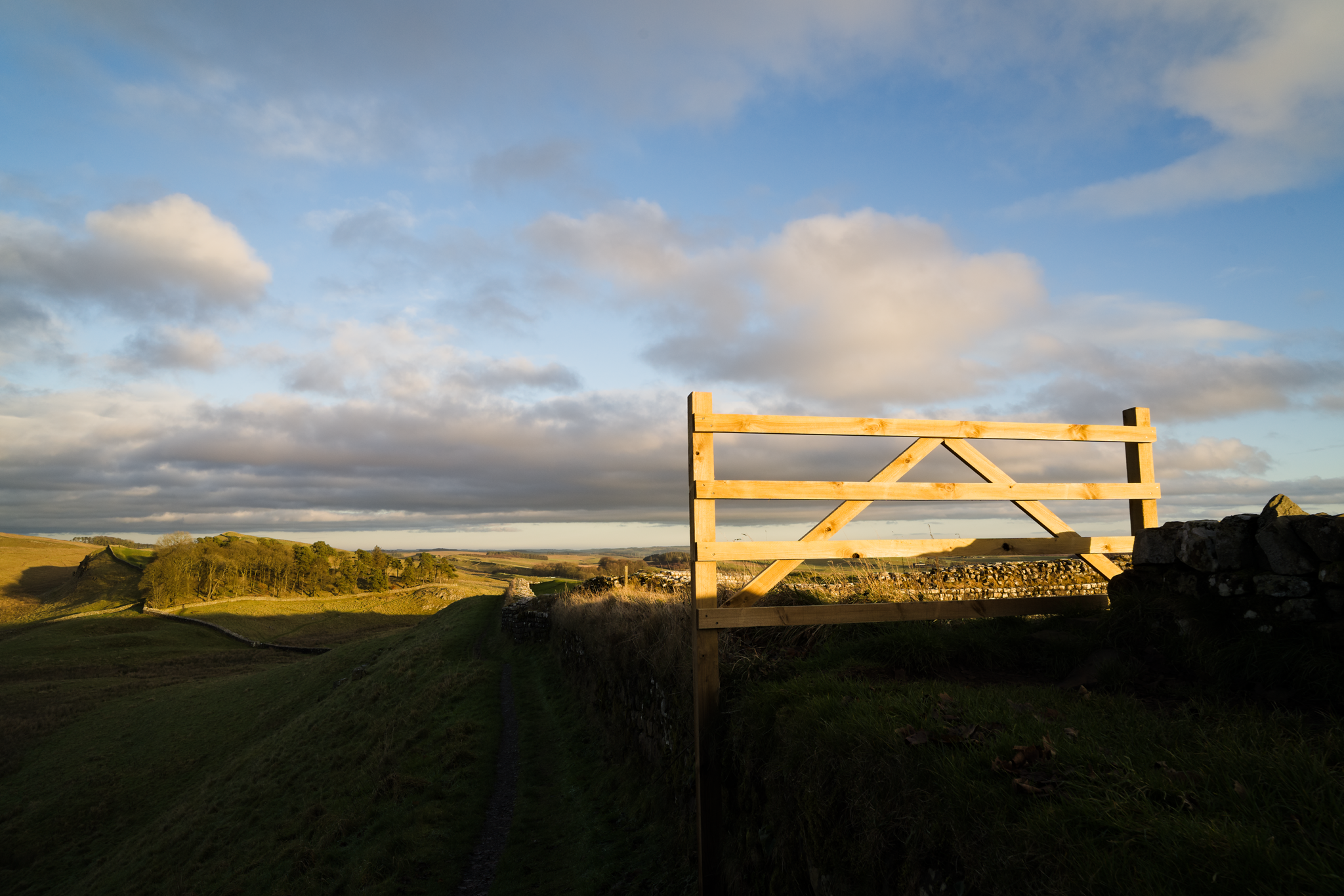 A section of fence, catching the low sun, blocks the path along the top of Hadrian's Wall. Countryside and light cloudy sky.