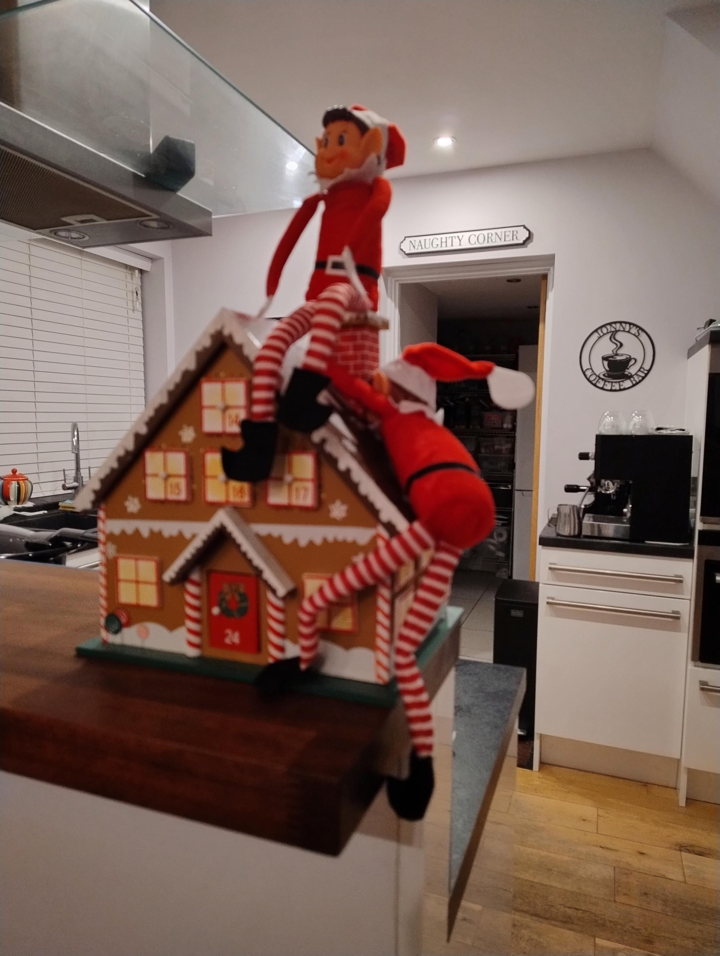Elf on a shelf character scrambling up a wooden advent calendar house. His companion is already seated on the chimney.