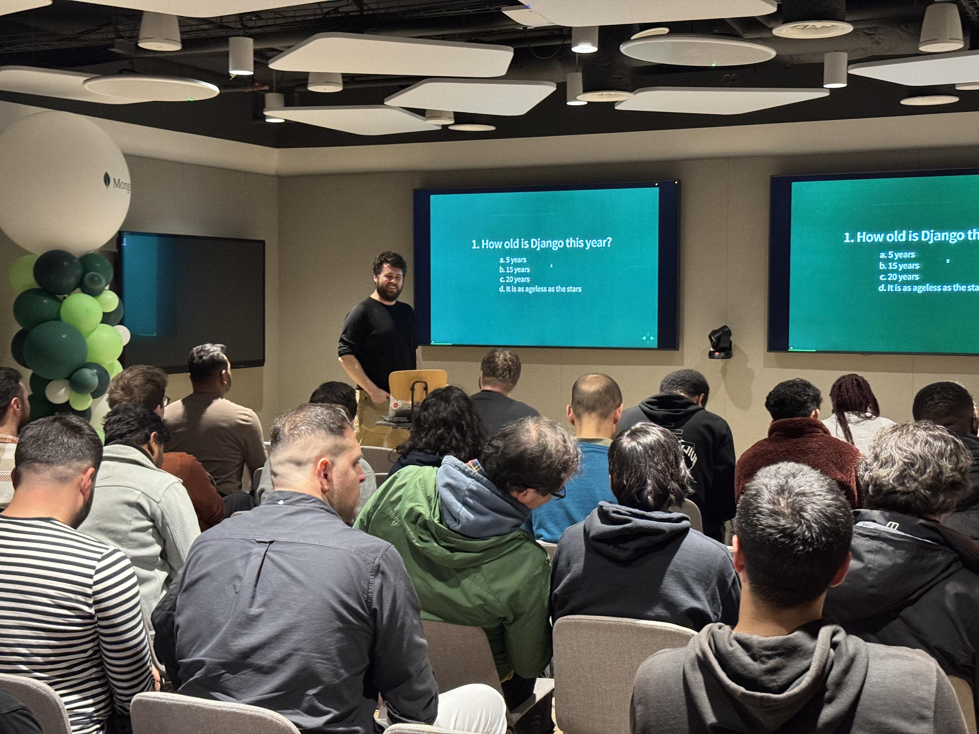 Adam Johnson hosting the Django Quiz at the Django London Meetup in the MongoDB event space, standing in front of quiz questions on large screens while attendees follow along from their seats.