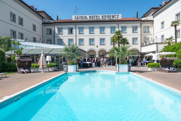 Photo of the Savoia Hotel Regency in Bologna, the venue of PyCon Italia. The image shows the hotel building with its name visible on the facade, a swimming pool in the foreground, a green garden with palm trees, and an outdoor bar area. The sky is clear and blue, and the scene is bright and sunny, taken during the PyCon Italia 2025 conference.
