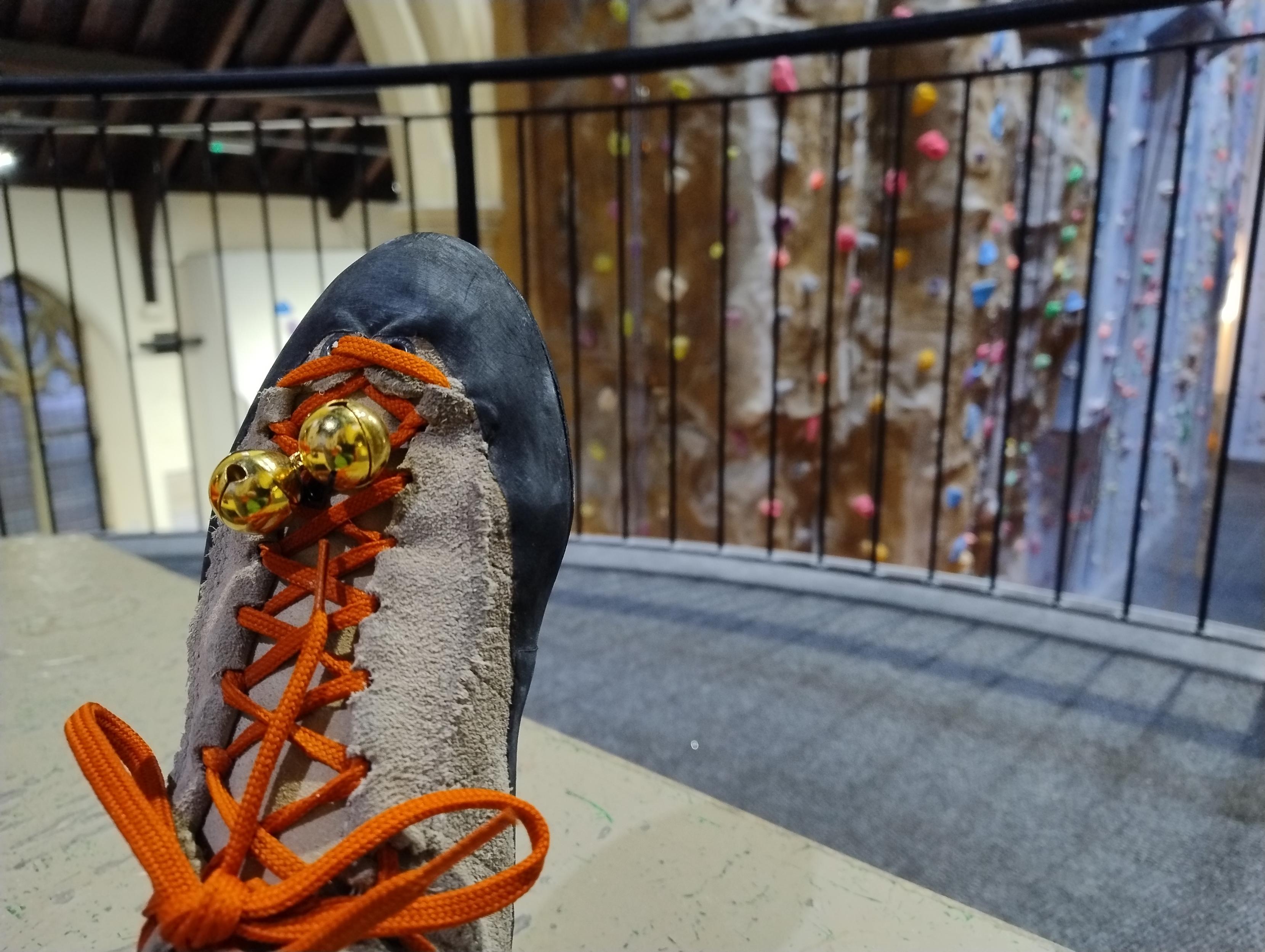 Rock climbing shoes with small Christmas bells tied to the laces. Indoor climbing wall in the background.