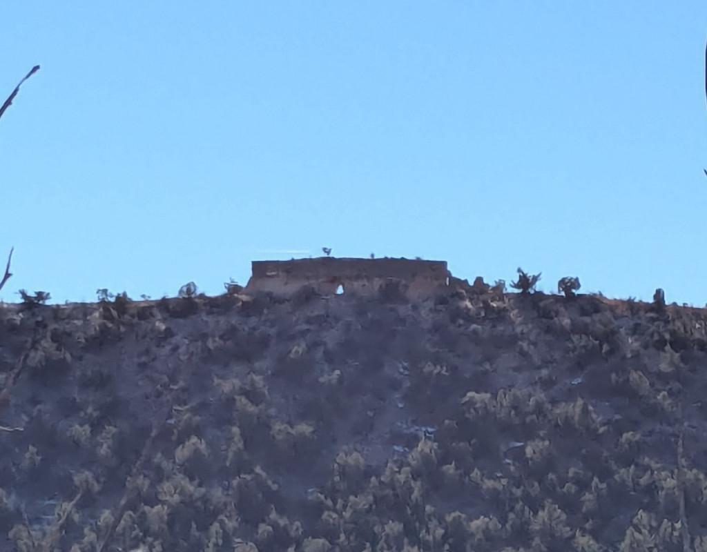 A photo of a rock arch taken from across a canyon. Standing on top of a ridge, the arch is a big rectangular flat-topped chunk of rock with a passageway through the bottom of it right in the center. It looks like it's made of two layers, a lighter one below and a darker one above. It has a small tree growing on its flat top. The ridge it's on is steep and covered with pinons and junipers.