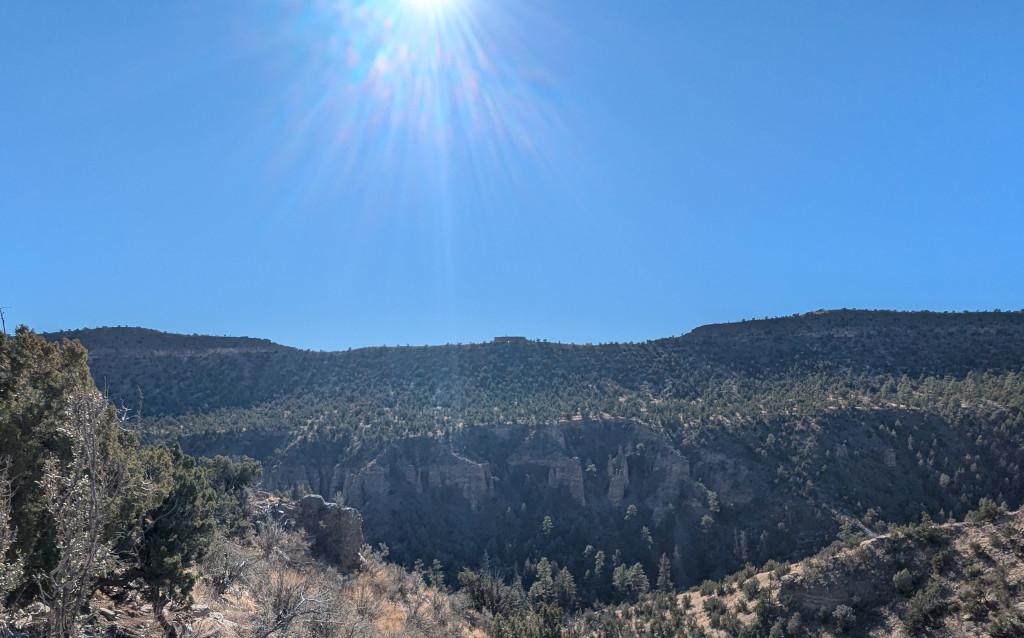 Looking across a canyon at a tree-covered ridge. On top of the ridge, centered in the photo, is a rectangular flat-topped rock where you can just barely see a sliver of blue sky through a hole in the bottom center. The ridge slopes down to a vertical cliff, where you can barely make out a collection of hoodoos or "tent rocks" on the face of the cliff. We're looking into the sun, which is just above the top of the photo, as you can tell from the colorful sun flare at the top. The sky is clear blue.