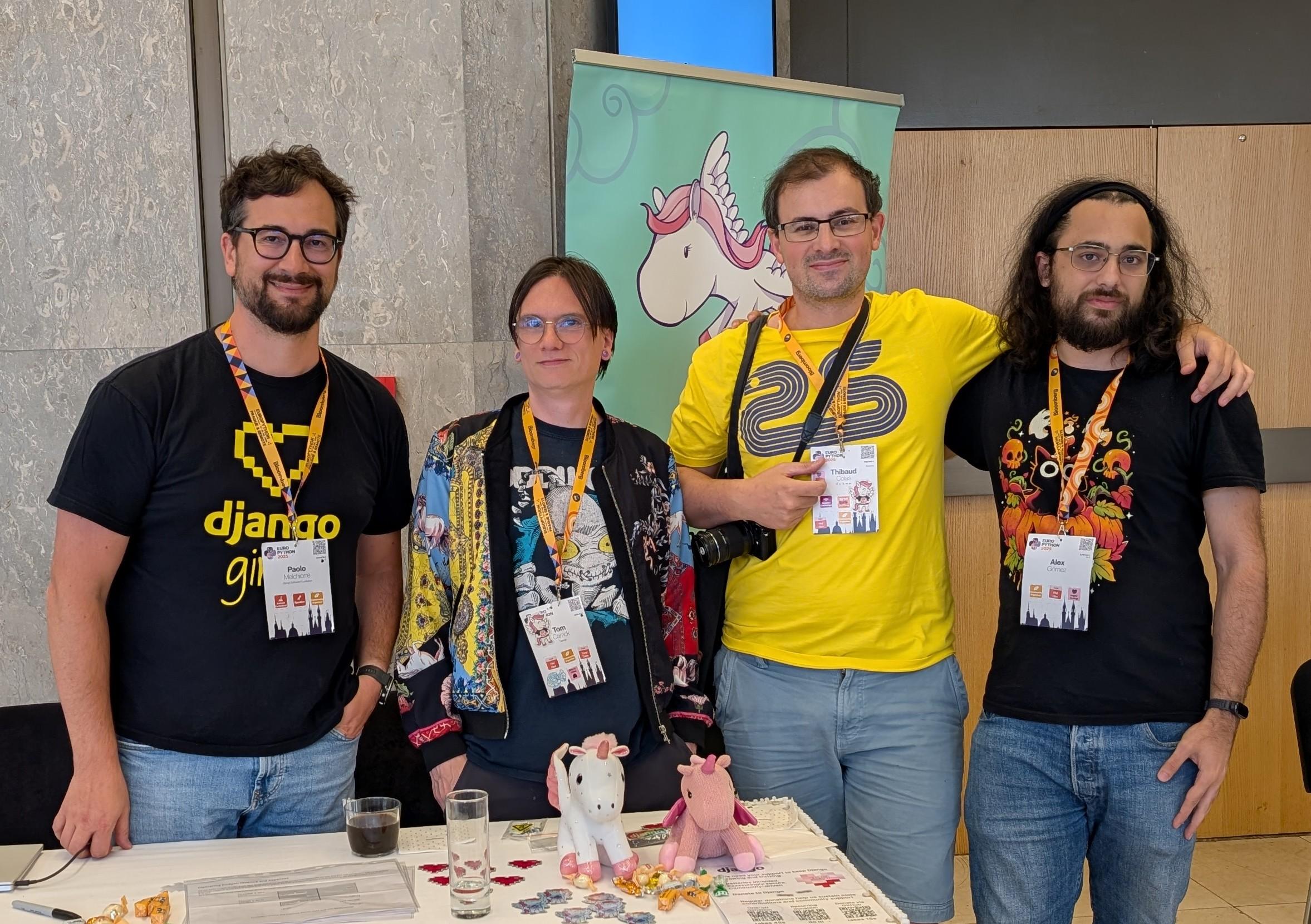 Group photo at the Django Software Foundation booth during EuroPython 2025. I am standing behind the table together with other DSF members. Django stickers are spread on the table and a large Django banner is visible behind us. Everyone is smiling, showing a relaxed but purposeful community moment focused on governance and support.