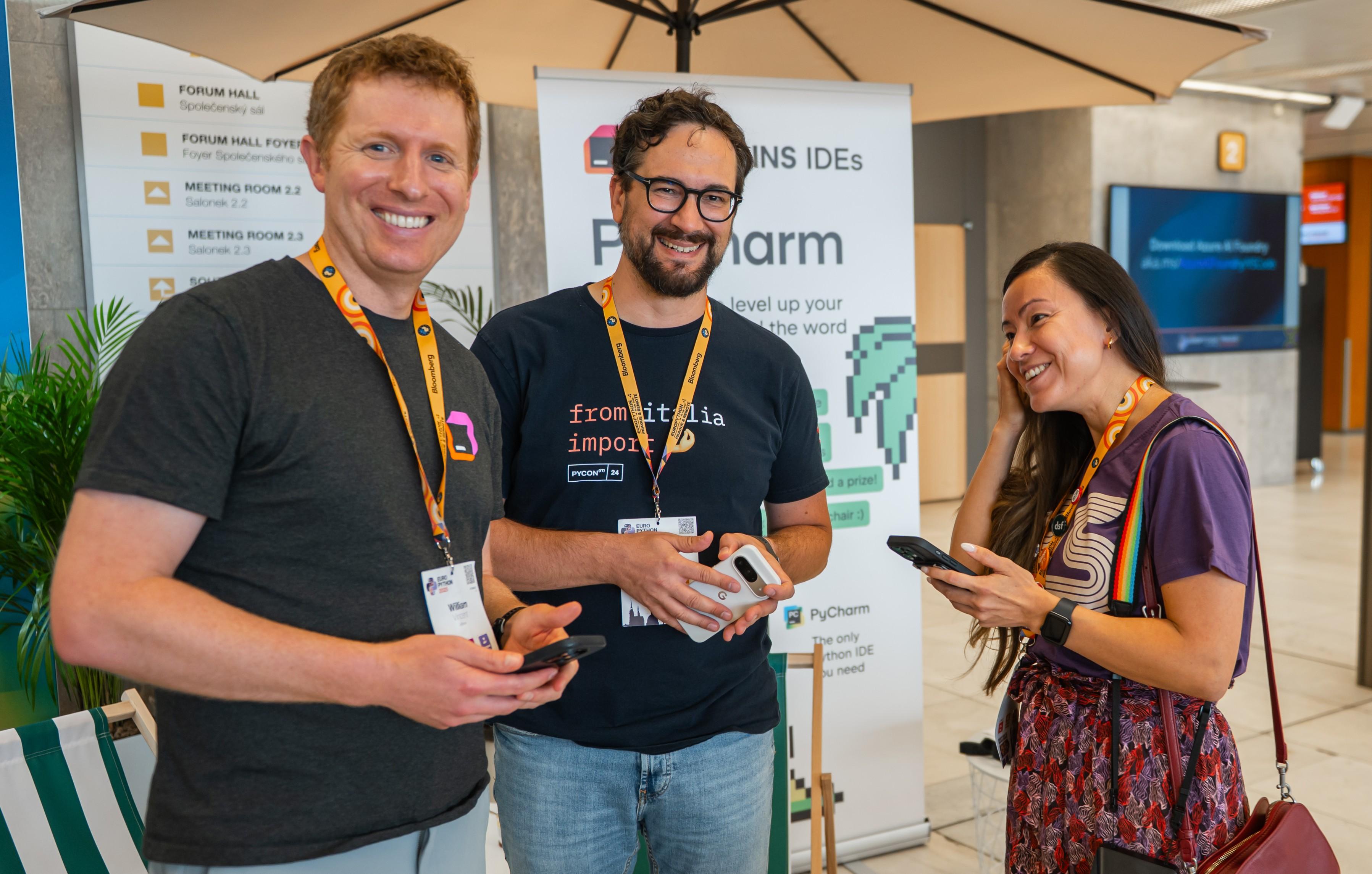 Photo taken in the conference hallway at EuroPython 2025 showing me standing and smiling at the camera, with Will standing to my left smiling at the camera, and Katia standing to my right smiling while looking at us and holding a phone.