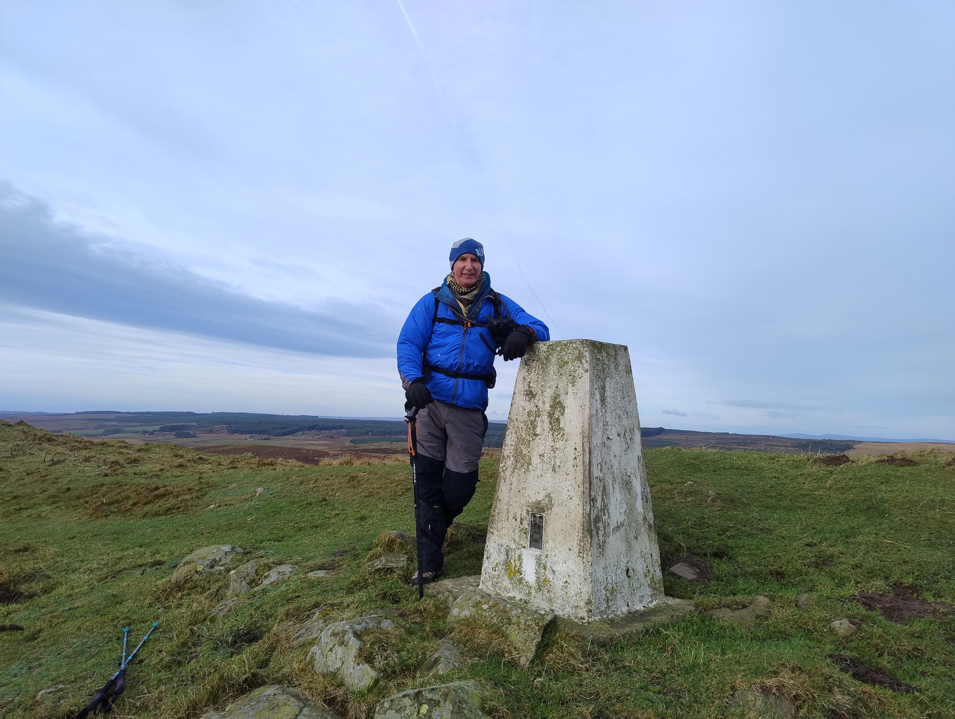 Hiker dude standing beside a white concrete trig point.