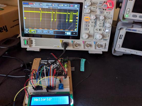 A Metro (Arduino) board connected by multiple colored wires to an LCD character display. The display reads "Hellorld!" Behind the circuit is an oscilloscope with a trace displaying some digital logic pulses.