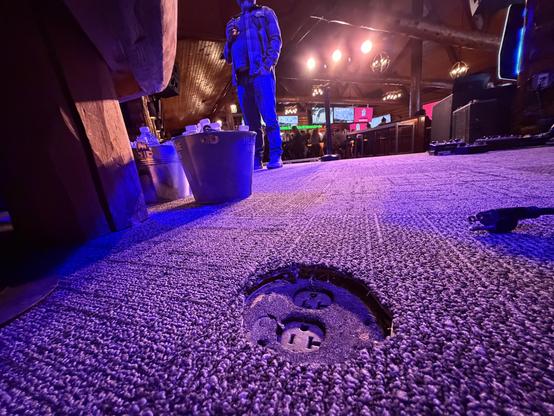 Een man in een donkere ruimte met een paarse vloer en een open putdeksel, met metalen emmers en een podium in de achtergrond.