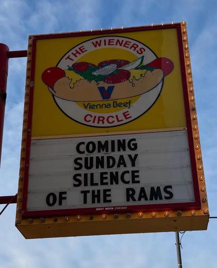 A yellow sign with a hot dog illustration advertises "The Wieners Circle" and a humorous event on a clear day.